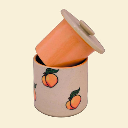 A speckled beige ceramic butter bell with a matching lid, decorated with a pattern of orange peaches with green leaves, set against a neutral light gray background.