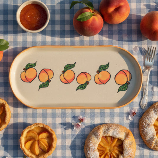An oval ceramic serving tray with a pale cream background, decorated with a row of hand-painted peaches and green leaves. The tray features a warm orange-brown rim and is photographed against a neutral light gray background.