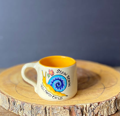 A hand-painted ceramic mug with a snail illustration and the lyrics 'Slow down you're doing fine' in a whimsical style.